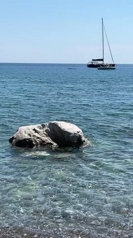 A serene seascape features a clear blue sky and calm, turquoise water. In the foreground, a large, grey rock breaks the surface near a pebble-strewn shoreline. Further out, a white sailboat with a tall mast dominates the horizon, accompanied by a smaller boat. Several people are visible in the water, swimming or enjoying the sea. The scene is peaceful and bathed in bright daylight, suggesting a pleasant day at the beach.