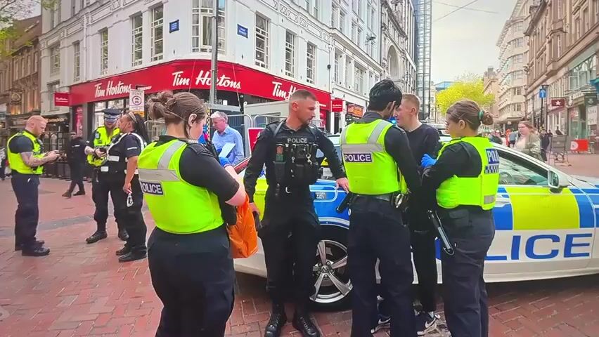 A group of police officers in uniform are gathered around a parked police car on a city street. Some officers are wearing high-visibility vests over their uniforms. One female officer is holding an orange plastic bag. A young man in a dark shirt and jeans is standing between two female officers. Another male officer is standing behind the young man, speaking to him. In the background, there are buildings, including a Tim Hortons coffee shop, and other pedestrians. The police car has the word "POLICE" written on its side. The scene appears to be a calm interaction between law enforcement and a civilian.