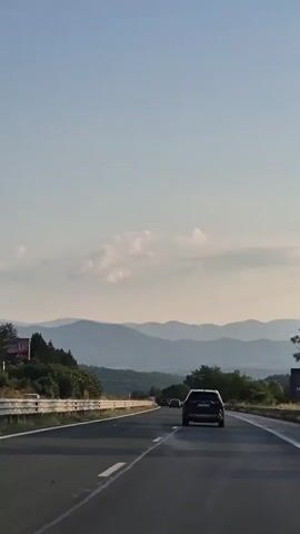 The video captures a scenic drive on a highway in Brestnitsa, Bulgaria, likely during the late afternoon or early evening due to the soft, warm lighting. The camera is positioned inside a vehicle, looking forward along the road.

The primary subjects are several cars traveling on the highway. In the foreground, a dark-colored SUV is prominent, with another similar vehicle ahead of it. Further down the road, a line of cars can be seen.

The setting is a rural highway bordered by a guardrail and lush green trees and vegetation on the left. In the distance, a series of rolling hills or mountains form a majestic backdrop, their layers fading into a hazy blue. The sky is a clear, pale blue with a few scattered, wispy clouds, suggesting pleasant weather.

The activity is simply driving, with the cars moving along the open road. The overall mood is peaceful and serene, enhanced by the natural beauty of the landscape and the gentle movement of the vehicle. There are no explicit emotions conveyed, but the imagery evokes a sense of calm travel and appreciation for the surroundings.

There is some text visible on the license plate of the closest SUV, which appears to read "EB 0871 BC". On the left side of the highway, a billboard is partially visible with text that is difficult to read clearly but seems to include the word "GORY".