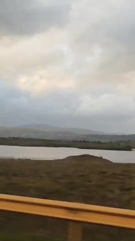 The video shows a view from a moving vehicle. A body of water is visible in the middle ground, with rolling hills and a cloudy sky in the background. The foreground features a blurry yellow railing. Trees are visible on the left side of the frame. The overall scene is overcast and appears to be a rural landscape.