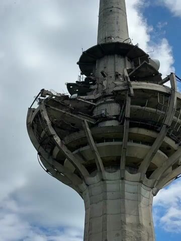The video shows a tall concrete tower, extensively damaged around its midsection. The damage appears to be structural, with sections missing and rebar exposed. The tower narrows as it ascends, with various antenna-like structures attached to its upper portions. The sky, with white clouds and patches of blue, forms the backdrop. The camera angle is looking upwards towards the tower.