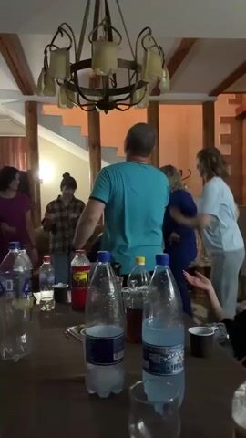 The video shows a group of people dancing and socializing around a table in what appears to be a rustic indoor setting with a chandelier and wooden beams. There are several bottles of drinks and cups on the table. The camera is positioned low, looking up at the table and the people.