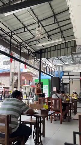 The video shows a scene inside a covered outdoor restaurant or food court. A white ceiling fan is mounted on the metal roof structure. Several tables with wooden chairs are set up. In the foreground, a man wearing a green and white striped shirt sits at a table, facing away from the camera. To his left is a vintage sewing machine base used as a table leg. Further back, a woman in a brown top and shorts stands near a green vending machine labeled "THAI BANK HERE" with a Visa logo. She appears to be interacting with it. Other patrons are seated at tables in the background, some facing away from the camera, engaged in eating or conversation. The environment suggests a casual dining setting, likely in Southeast Asia due to the architectural style and the presence of a Thai bank machine. The lighting appears to be natural light supplemented by overhead fluorescent lights.