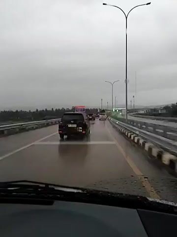 The video shows a view from inside a vehicle driving on a wet highway. Raindrops are visible on the windshield, and the windshield wipers are intermittently clearing the view. Several cars are driving on the highway ahead, with a dark SUV being the most prominent. The sky is overcast and gray. A dark plume or column is visible rising into the sky in the distance, to the left of the highway. The plume appears to be moving and is a prominent feature in the sky. There are roadside barriers and streetlights along the highway. The overall impression is of driving on a highway in inclement weather, with an unusual atmospheric phenomenon occurring in the distance.