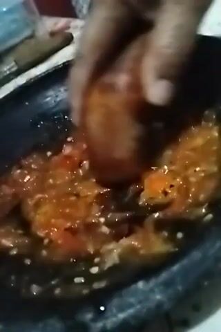 A close-up shot shows a person's hand holding a wooden pestle and grinding red ingredients in a dark stone mortar. The ingredients appear to be tomatoes and possibly chili peppers, with some white specks possibly indicating garlic or onions. The texture is coarse and liquid is visible. The scene is dimly lit, suggesting an indoor setting.