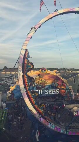 The media file captures an exhilarating scene at a bustling carnival or fair, possibly in Gloucester, USA. The primary focus is a large, circular amusement ride adorned with vibrant lights and a nautical-themed mural depicting pirate ships and turbulent seas. The words "SEVEN SEAS" are prominently displayed on the ride's central panel.

Numerous people are visible, gathered below and on the ride, suggesting it is in operation. The ride itself appears to be a thrilling pendulum or swing attraction that rotates riders in a large arc. The weather appears pleasant with a partly cloudy sky, hinting at late afternoon or early evening. The presence of buildings and what seems to be a marina in the background provides a strong sense of location. The overall mood is energetic and festive, characteristic of a fairground atmosphere. Another ride, labeled "FIREBALL," is also visible in the foreground, further emphasizing the carnival setting. American flags flutter at the top of the large circular ride, adding a patriotic touch.