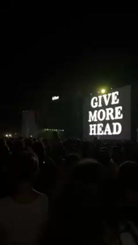 From a low-angle, partially obstructed viewpoint, the video captures a concert with a large crowd. The scene is dark, with illumination primarily from the stage lighting and large video screens displaying a performer. The focus is on the stage and the performer shown on the screens, with the crowd appearing as a mass of silhouettes. People are likely standing or moving, as one person is seen partially turning their head towards the left. The environment suggests an outdoor venue, with the stage as the focal point of activity.