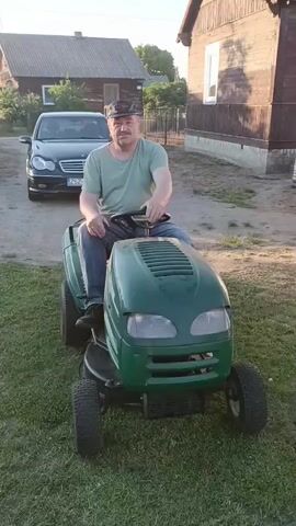 An older man wearing a green t-shirt, blue jeans, and a black baseball cap sits on a green riding lawn mower. He is in a grassy yard with a black car parked behind him and two wooden houses in the background. He gestures with his hands and appears to be talking or singing. The video is 4 seconds long.