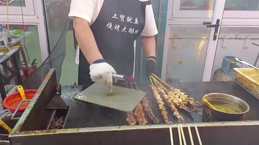 The video shows a person wearing a white t-shirt, a black apron with Chinese characters, and white gloves, standing behind a flat-top grill. The person is holding a metal tool and appears to be pressing down on something on the grill. There are several skewers of food on the grill, some of which appear to be meat and seafood. To the left of the grill, there is a red container and a bottle of yellow liquid. To the right of the grill, there is a bucket of liquid and some yellow-topped containers. In the background, there is a window with a view of what appears to be a restaurant or food stall. The lighting in the video is artificial, and there are some colorful lights visible in the background. The overall impression is that of a food preparation area, likely a street food stall or a casual eatery.