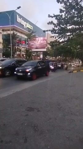 The video shows a street scene with traffic. In the background, a large building with "living Plaza" signage is visible. Several cars and motorcycles are on the road, and some are moving. The lighting suggests it might be dusk or early morning. A black SUV is in the foreground, and a white car is behind it. A red scooter is also visible. The road appears to be asphalt. The overall impression is a busy urban street.