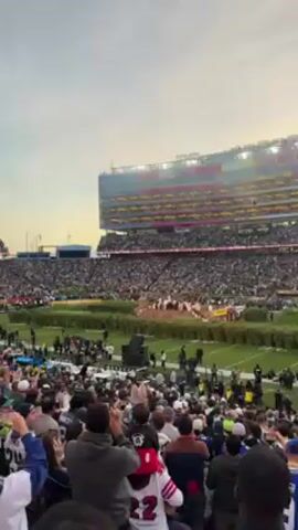 The video shows a stadium filled with a large crowd of people. The event appears to be a large sporting event or concert, as there is a stage set up on the field. There are large video screens on either end of the stadium. The camera is positioned among the crowd, looking towards the field. The sky is overcast. The stadium appears to be Levi's Stadium.