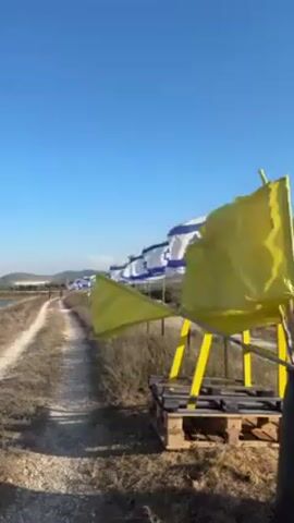 The video depicts a row of Israeli flags waving in the wind alongside a dirt path. To the right of the path, there is a small platform with yellow flags also waving. The sky is clear and blue, with distant hills visible on the horizon. A single figure can be seen walking along the path in the distance.  The foreground is filled with dry grass and vegetation.