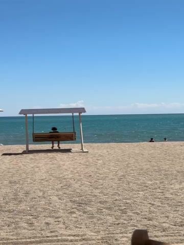 A person sits alone on a wooden swing on a sandy beach. The swing is under a white canopy supported by two white posts. The beach is made of light brown sand. In the background, there is a body of water with gentle waves. The water is a turquoise color and meets a clear blue sky at the horizon. There are faint white clouds visible near the horizon. Two other people are partially visible in the water in the distance. The sun is shining brightly, casting shadows. The scene is peaceful and serene.