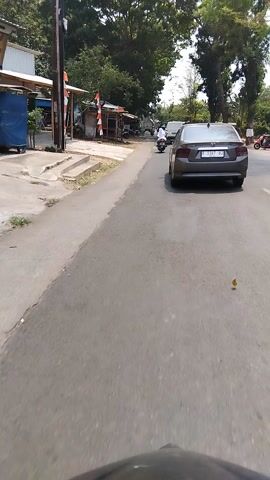 The video captures a view from a moving vehicle, likely a motorcycle, on a paved road in Pasekaran, Indonesia. The scene is a daytime street view with lush green trees lining the left side of the road and a few buildings visible in the background. The weather appears to be sunny, as indicated by the bright lighting and the clear sky.

The primary subjects are vehicles on the road. A silver sedan is prominently featured, driving ahead of the camera. A motorcycle rider, wearing an orange jacket and a white helmet, is also visible. The rider is on a white scooter, and the orange jacket has the word "Food" printed on the back, suggesting they might be a food delivery person. There are other motorcycles and some parked vehicles further down the road.

The activity is the progression of traffic on a road. The camera vehicle is moving, following the silver car and passing other vehicles. On the left side of the road, there are some small stalls or shops with people around them, and what appears to be a crossing gate, suggesting the road might be near a railway crossing.

Notable details include the license plate of the silver car, which appears to read "C 1167 BD". The time of day is clearly daytime, and the overall mood is that of ordinary daily activity. The location cues like the vegetation, the style of buildings, and the appearance of the vehicles point to a Southeast Asian setting, consistent with the provided location context of Pasekaran, Indonesia.
