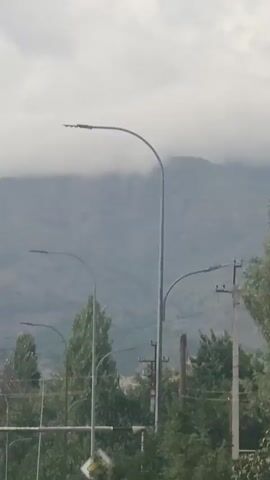 The video shows a street with multiple streetlights and trees. In the background, there are mountains covered in fog or clouds. The sky is overcast. There is a power pole with wires visible in the middle of the frame. There is also a yellow and white road sign visible. The camera appears to be moving, likely from a vehicle, as evidenced by the slight shake and the way the scene unfolds.