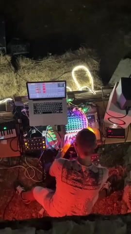 A person sits in front of a table filled with electronic musical equipment. The person is viewed from behind and is wearing a white shirt with a patterned design. They are wearing light-colored pants and white sneakers. The table has a laptop, several modular synthesizers, a midi keyboard, a drum machine, and speakers. The equipment is connected with numerous cables and is illuminated by colorful LED lights. The person's hands are on the drum machine, interacting with its illuminated buttons. The environment appears to be outdoors at night, with hay bales visible in the background and a glowing string of lights. The scene is lit with red and orange tones, casting a warm glow on the person and the equipment.