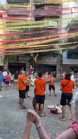 The video shows an outdoor street scene with a band playing music. The band members are wearing orange shirts and shorts. They are playing various instruments including trumpets, a tuba, and a double bass. In the background, there are buildings decorated with colorful banners. Several people are standing and watching the band, some are holding hands and dancing. The ground is covered with confetti.