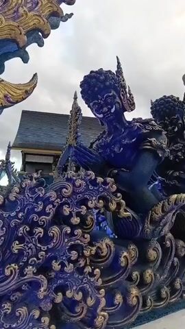 The video shows a close-up view of a blue and gold artistic sculpture. The sculpture depicts several figures with dark blue skin and ornate headdresses. The figures are in a seated or kneeling position, with their hands pressed together in front of them. The sculpture is set against a background of a cloudy sky and a building with a dark tiled roof. The overall impression is of a religious or ceremonial artwork, likely from Southeast Asia given the style and ornamentation. The lighting is somewhat dim, suggesting it may be overcast or early morning/late evening. The camera slowly pans across the sculpture, highlighting the intricate details of the carvings and the vibrant colors.
