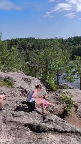 Two young women are sitting on a rocky cliff overlooking a lake. The woman on the left has blonde hair and is wearing a white tank top and black shorts. The woman on the right has brown hair and is wearing a pink and white checkered top and shorts. She is holding a phone and appears to be showing something to the other woman. The lake is surrounded by a dense forest of green trees. The sky is blue with some white clouds. The overall scene is peaceful and idyllic, suggesting a recreational activity like hiking or enjoying a view.