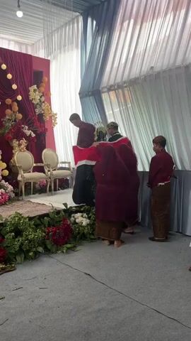 A wedding ceremony is taking place. The bride and groom are standing on a stage with floral decorations. A red and white cloth is being held up, possibly as part of a ritual. There are several people in traditional attire. The environment appears to be an indoor venue with draped curtains.