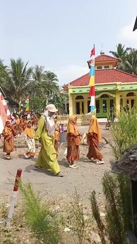 The video captures a procession of people, predominantly children dressed in traditional Muslim attire, walking along a dirt road. The children are seen in groups, some wearing orange and brown outfits, while others have on lighter-colored dresses. A few adults, also in traditional attire, are interspersed within the procession, guiding and accompanying the children. Prominently featured are several flags, including one large red and white flag that arches over the road, and numerous smaller flags in various colors, including red, white, blue, and yellow, strung along poles. In the background, a mosque with a red roof and yellow walls is visible, alongside some houses and palm trees. The environment appears to be a rural or semi-rural setting, with dry earth and vegetation. The overall impression is that of a community event or celebration.