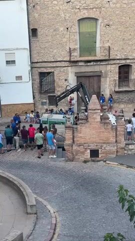 A group of people are gathered in a cobblestone square in front of an old stone building. An excavator is positioned in front of the building's entrance, with its arm extended towards a structure made of bricks and a green, possibly artificial, foliage. Some individuals are standing near the excavator, while others are observing from a distance. The building has multiple windows and balconies, and a large wooden door is visible behind the excavator. The scene appears to be taking place outdoors during the day, with a white building to the left and a curved wall in the foreground.