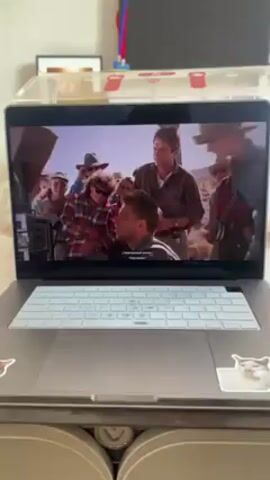 A laptop sits on a surface and displays a movie scene. Several people surround a man speaking into a microphone in a dusty, outdoor setting. The laptop has two cat stickers. The laptop sits under a clear plastic box. There is a red and blue object in the background.