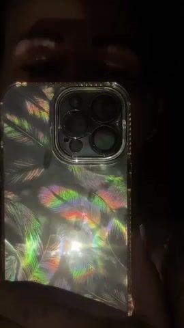 The video shows a close-up view of a person's face and an iPhone. The iPhone has a clear case with a holographic feather pattern. The camera is angled towards the user, taking a selfie in a dimly lit environment. The user's eyes and eyebrows are visible above the phone. The phone's camera module is prominent, reflecting light.