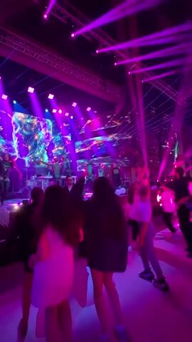 A concert is taking place on a stage in a nightclub. People are dancing in front of the stage, lit by purple and blue lights. The stage features a large screen displaying abstract imagery, and several performers are visible on stage. The camera is positioned behind a group of dancers, capturing their movements as they enjoy the music.