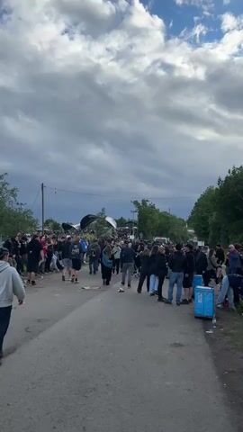 A large crowd of people, primarily young adults, is gathered outdoors on a paved road that appears to be part of an event or festival ground. The road is somewhat littered with discarded items like paper and plastic. On the far right, a blue portable bin or cooler is visible next to the road. Many individuals are walking in various directions, with a general flow towards the background where a large, possibly tent-like structure or stage is partially visible under an overcast sky with hints of blue. Trees line the distant edges of the area, and power lines traverse the scene overhead. People are casually dressed and engaged in walking, standing, and conversing in small groups.