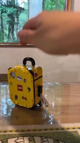 The video shows a person's hand interacting with a yellow Lego suitcase. The suitcase is decorated with various stickers, including the Lego logo, a cactus, and a heart. A luggage tag that reads "BILL" is attached to the handle. The hand pulls the handle upwards, extending the suitcase's telescopic handle and tilting the suitcase. The background appears to be a room with a wooden floor and a window looking out onto a forest. The person is manipulating the Lego suitcase as if they are preparing to travel with it.