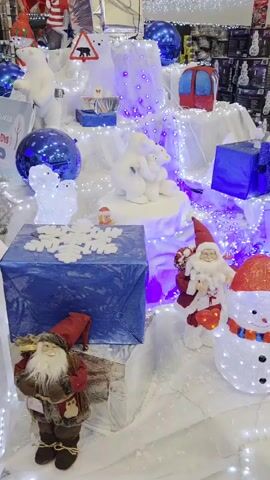 The video shows a Christmas display in a store. The display features white polar bears, a snowman, and a Santa Claus figure, all illuminated with white lights. There are also blue ornaments, blue gift boxes, and a red ski gondola decoration.  A reindeer figure is also visible. The scene is set against a backdrop of white fabric resembling snow, and blue and white Christmas lights illuminate the entire display. Shelves and boxed merchandise are visible in the background.