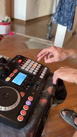 The video shows a close-up of a person's hands operating a DJ controller. The controller is black with a variety of buttons, knobs, and a small screen. The person's hands are positioned over the buttons and knobs, with fingers pressing and adjusting them. The background of the video is blurred but appears to be an indoor setting with tiled flooring and some furniture. The lighting is natural, suggesting it might be daytime. The overall impression is of someone actively engaged in using the DJ equipment.