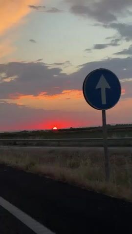 The video shows a sunset viewed from a moving vehicle. The sun, a vibrant red orb, is low on the horizon, partially obscured by a thin line of dark vegetation. The sky above is a gradient of colors, transitioning from pale blue at the top to shades of orange and pink near the sun. Scattered clouds, tinged with the same warm hues, drift across the sky. In the foreground, a dark asphalt road curves to the left, with white lane markings visible. To the right of the road, a metal guardrail separates it from a grassy field. A few thin, vertical posts are visible in the field, with a chain-link fence behind them. A utility pole with wires is also visible in the distance to the left. A triangular yield sign is present on the right side of the road.
