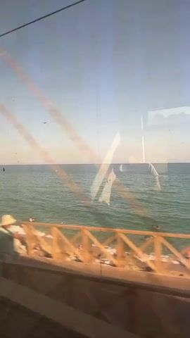 This media file captures a view of the sea from a moving perspective, likely a train or bus, given the slight blur and the roadbed visible in the foreground. The scene is set on a sunny day at Montgat, Spain, with the clear blue sky meeting the calm, turquoise sea at the horizon. A rocky shoreline, typical of the Catalan coast, separates the water from the unseen land.

In the water, a few small yellow buoys are visible, and a person is swimming, indicated by an orange object near them. The water is serene with gentle waves lapping the shore. Reflections on the glass obscure parts of the scene, showing a blurry silhouette of a person or object inside the vehicle. These reflections, along with the slight movement of the scene, suggest a journey is in progress. The overall mood is peaceful and relaxed, evoking a sense of travel and seaside leisure. No visible text is present in the media file.