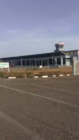 The video shows an airport building with an air traffic control tower. In the foreground is a parking lot with white stripes, and a sign indicating the entrance to the airport. A person on a motorcycle can be seen driving past the entrance sign towards the airport building. The sky is blue and the environment appears to be sunny. The ground around the parking lot is covered in brown vegetation.