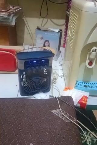 The video shows a portable air cooler fan in operation. The fan is black and blue with a mesh front. The top of the fan has blue indicator lights. The fan is plugged in and positioned on the floor. Next to the fan is a white tissue box with red and orange branding. To the right of the fan is a tall, cream-colored water dispenser. Behind the fan is a box with a picture of a woman on it, labeled "AIR COOLER FAN". To the left of the fan is a red pouch or bag. The floor is tiled. The scene appears to be indoors, possibly a room in a house.