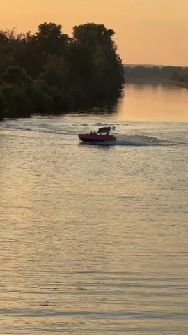 A motorboat with several people on board speeds across a body of water, kicking up a wake. The boat is red and white and has a canopy. The surrounding environment appears to be a river or large lake with trees lining the shore in the background. The sun is setting, casting a warm, golden light on the water and sky. The video appears to be taken from a slightly elevated position, looking down at the boat and the water. The boat is moving from left to right across the frame.