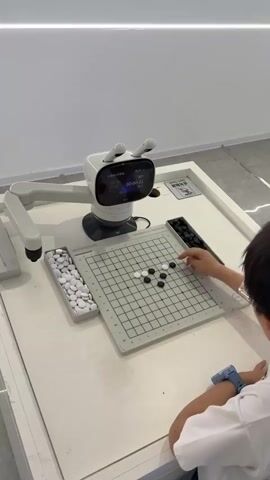 The video shows a young person playing the game Go against a robot. The game is set up on a table with a gray grid board. The player sits on the right side of the table, wearing a blue watch. They are placing black stones on the board. Opposite them is a white robot with a small display screen showing a timer. The robot has a robotic arm extended towards the game board, poised to make a move. There are containers for white and black stones on either side of the board. The table is white and the background is a plain white wall.