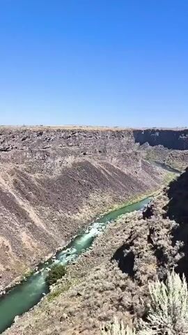 The video shows a drone view of a canyon with a river flowing through it. The canyon walls are steep and rocky, with sparse vegetation. The river is a bright green color and appears to be flowing rapidly in some sections. There is a man-made structure, possibly a dam or bridge, spanning the river. The drone flies over this structure, offering a closer view of its construction. The video ends with a shot of the river flowing away from the structure into the distance, with the canyon walls continuing to flank it. The sky is clear and blue, indicating a sunny day. The overall impression is one of a natural landscape with human intervention.