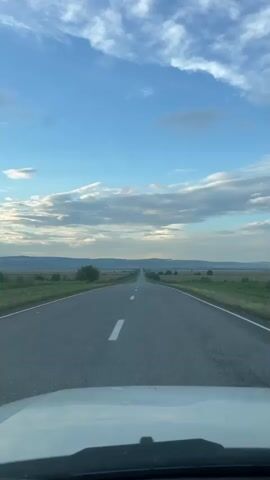 The video captures a view from a vehicle driving on a straight, empty asphalt road in Khumalag, Russia. The road is lined with white markings and extends towards the horizon, disappearing into a landscape of rolling hills and fields. A solitary tree stands on the left side of the road, adding a touch of greenery to the otherwise agricultural scenery.

The sky is a vibrant blue, adorned with scattered white clouds that suggest fair weather. The sun appears to be either rising or setting, casting a soft light and creating a serene atmosphere. The overall mood is one of peaceful travel and open exploration, with the long, uninterrupted road symbolizing a journey forward. There is no visible text in the media.