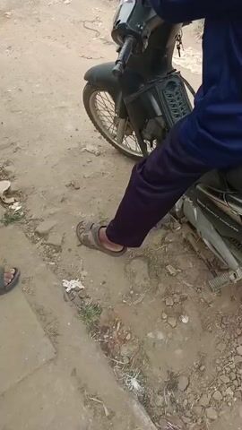 The video shows a person sitting on a motorcycle. The motorcycle is dark colored with a red rim. The person is wearing dark pants and sandals. The ground is dirt and gravel. Another person wearing black sandals is visible in the lower left of the screen. The motorcycle is on a dirt path next to a paved road. The paved road has grass growing along its edge. There are buildings visible in the distance. The motorcycle then moves down the paved road. The person on the motorcycle is wearing a blue jacket. The motorcycle is passing by a black SUV and then drives away from the camera.
