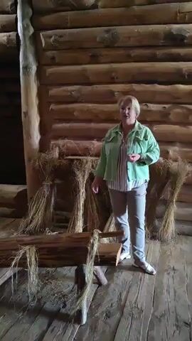 A woman in a green jacket and grey pants stands in a rustic wooden structure, likely a barn or historical display. She is facing the camera and gesturing with her right hand. Behind her, bundles of dried stalks are propped against a log wall, and in front of her, a wooden contraption with a log and what appears to be a scythe-like blade is visible. The floor is made of wooden planks. The woman's blonde hair is short and styled. She appears to be explaining something about the objects in the scene.