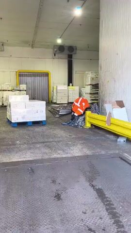 The video shows a forklift in a warehouse setting.

A worker in an orange safety vest is seen near a yellow barricade on the right side of the frame. Further back, a blue pallet loaded with white boxes sits in the middle of the warehouse floor, with rows of similar boxes stacked against the back wall. The warehouse is well-lit, with several ventilation systems visible along the ceiling. The floor is metal and textured, and a large metal plate is in the foreground. The forklift, painted in a faded teal color, is parked on the left side of the frame, facing toward the center.