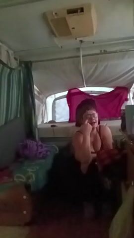 A woman with dark hair, glasses, and a blue sports bra is sitting inside a pop-up camper. She is looking at her phone. A bed with a red shirt on it is visible behind her, as is an air conditioner mounted on the ceiling. There is a striped curtain on the left, and several pillows and pieces of cloth are nearby.