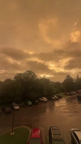 The video captures a view from above looking down on a parking lot and surrounding trees. The sky is overcast with dramatic, orange-tinted clouds, suggesting either sunset, sunrise, or smoke from a wildfire. Sunlight breaks through the clouds in some areas, creating bright patches. The parking lot is wet, indicating recent rain, and several cars are parked there, including a red car in the foreground, a dark SUV, and others further back in various colors. Trees with dense green foliage form a backdrop to the parking lot. The overall atmosphere is moody and somewhat dramatic due to the unusual sky coloration and weather conditions.