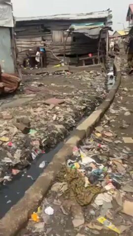 The video depicts a poor, densely populated area characterized by dilapidated wooden structures and widespread litter. A narrow, open concrete drainage ditch runs through the center, filled with dark water and waste. Debris and trash cover the ground, creating an unsanitary environment. Several people are visible: a woman stands on a raised wooden platform outside a building, and a child is seen playing nearby. Overall, the scene portrays a setting of poverty and poor sanitation.