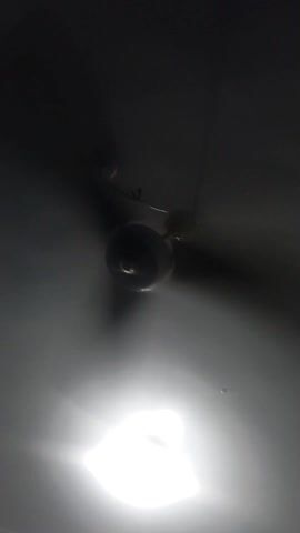 A ceiling fan spins slowly beneath a bright, square-shaped light fixture. The room is otherwise dimly lit, with the fan and light being the primary objects visible. The camera is pointed directly upwards.