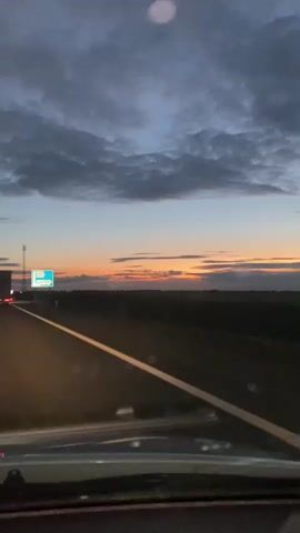 A truck is driving on a highway at dusk. The sky is cloudy with hints of orange and pink from the setting sun. There are streetlights and other vehicles on the road. The camera appears to be inside another vehicle, looking out the windshield.