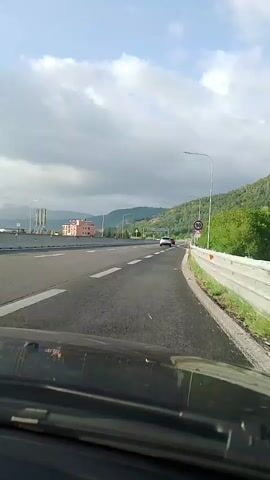 A first-person perspective video shows a view from the dashboard of a car traveling on a highway. The highway has multiple lanes and is bordered by a concrete barrier and a grassy embankment. Speed limit signs indicating 90 km/h are visible on the right side of the road. In the distance, there are hills covered with trees, and a building with a pink facade is visible on the left. The sky is cloudy, with patches of blue visible. Two cars are ahead in the distance, one white and one red, traveling in the same direction.